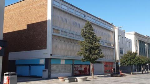 A large building with its shutters closed and boarded up. There are trees in front of building.