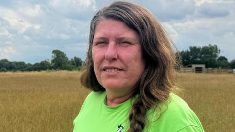 A woman with braided hair wears a lime green t-shirt and stands in a field of hay.