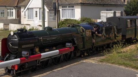 A steam locomotive about a third of the size of a regular locomotive. It is dark green and pictured on a level crossing. Bungalows can be seen in the background.