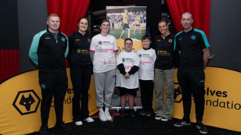 Wolves women players Anna Morphet and Tammi George with children and volunteers at the Wolves Foundation, standing in front of a display that features a picture of Wolves players during a match.