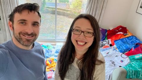 A man and a women taking a selfie with loads of bright coloured football shirts in the background.