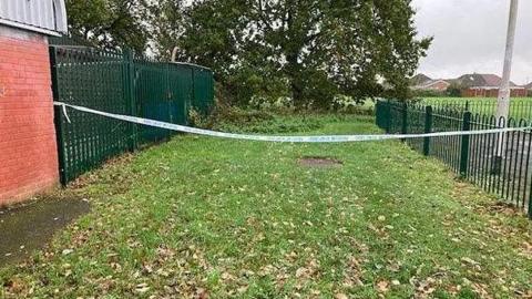 Police tape cordons off an area of grass spotted with leaves, with the tape attached to green metal railings at either side.