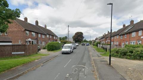 A road with houses on either side. Cars parked on both sides of the road.