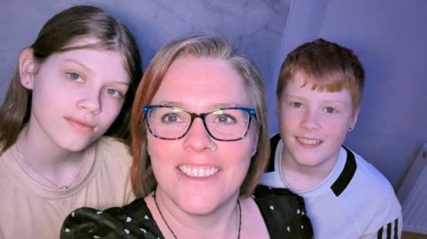 Shows a girl on the left, a woman in the middle wearing glasses with a ring through her left nostril, and a boy with red hair on the right