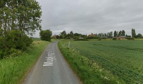 Mucking Hall Road, a narrow country lane with no hedges and fields on either side of the single-lane road