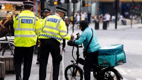 Two police officers in hi-vis can be seen at the junction of Tottenham Court Road and Oxford Street. One is seen speaking to what appears to be a Deliveroo driver on an e-bike.
