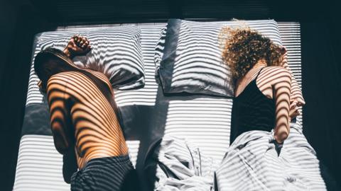 A man and woman lay next to each other in a double bed. Sun is peaking through blinds creating a striped pattern on them and the bed.