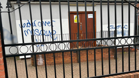 A white building with brown double doors behind black iron gates. On the wall, sprayed painted graffiti reads "undercover mosque" and "Islamic" with a word underneath it blurred out