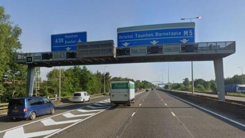 Junction 29 of the M5 in Exeter on a sunny day. Several cars, vans and lorries are on the road. There are four lanes on the road. One is a filter lane for people coming off at the junction. A gantry with blue signs for the A30 Honiton and Exeter Airport and the M5 up to Bristol, Taunton and Barnstaple on the road.