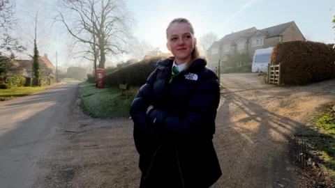 Ava, aged 14, stands in a country lane watching out for her taxi.  She is wearing her school uniform and has a bag over her shoulder.