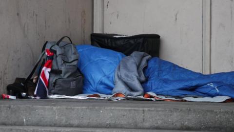 The shape of a person can be seen laying in a blue bin bag in a doorway. They have a grey rucksack with a Union Jack flag tied on it.
