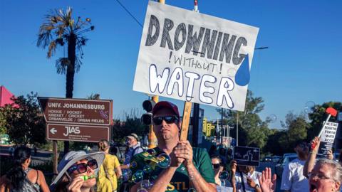 A man wearing sunglsses, baseball cap and green sports shirt, holds up a plard that says 'Drowning without Water' in Johannesburg in February 2025
