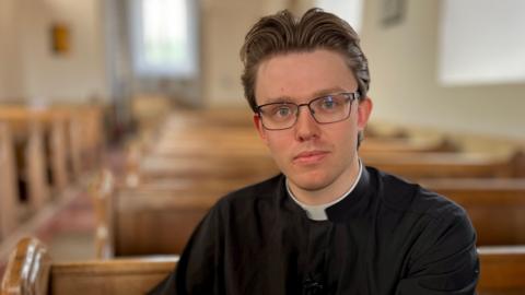 The Reverend Dr Edmund Smith is sat on a wooden pew, with rows of wooden pews behind him. He is looking directly at the camera and is wearing glasses.