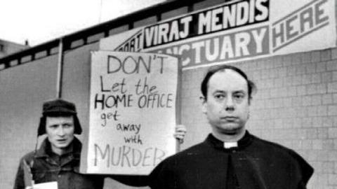 Father John Methuen is standing in front of the Church of the Ascension in Manchester. On the building are the words: "Viraj Mendis sanctuary here."
Behind Father Methuen is a man holding a sign which reads "Don't Let the Home Office get away with MURDER".