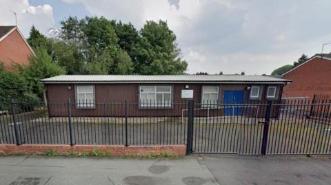 A single-storey building with a black fence separating an exercise yard from the pavement. The building has a set of blue double doors and a series of windows and wooden cladding.