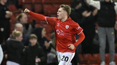 Charlie Lennon runs on the pitch with his right arm held in the air and fist clenched in celebration after scoring a goal. He is wearing Barnsley kit