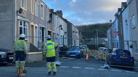 Two workmen in hi-vis jackets stand on Hugh Street, which is partly fenced off. Damage to a house roof can be seen on the left side of the street. A number of cars are parked on either side of the road.