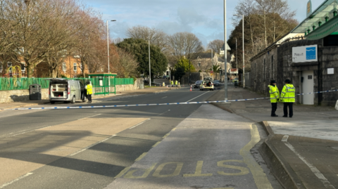 A police road closure - there are two officers stood on the pavement in front of police tape and there is a police car within the cordon further down the road