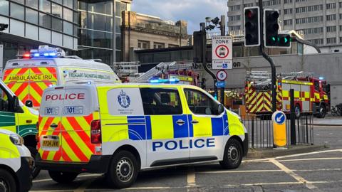 A police van, ambulance and fire engine stopped beside some traffic lights beside some city buildings