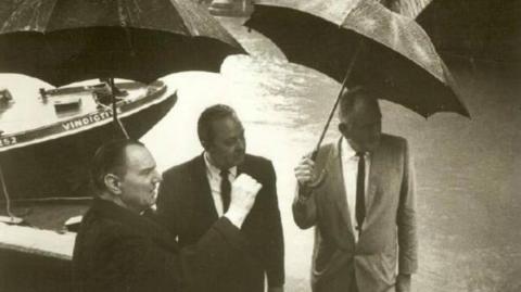 A black and white photograph showing Robert McCulloch (right) with his business partner C.V. Wood (centre), standing under umbrellas on a path by the River Thames and looking into the distance