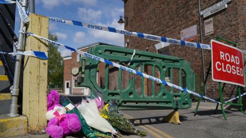 Flowers left at the scene of the stabbing. The are placed in front of green barriers with blue and white police tape across the road and a red an white Road Closed sign