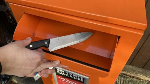 A close-up of a person holding a knife by its handle and placing it inside an orange bin.