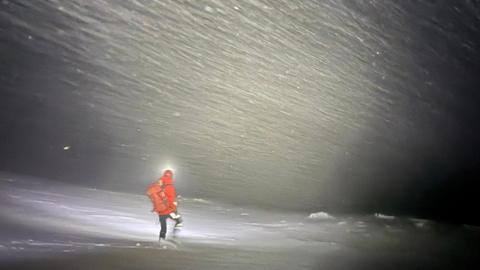 A rescuer wearing a red jacket, black jacket, walking boots and wearing a helmet and rucksack is walking through a blizzard while illuminated by torchlight.