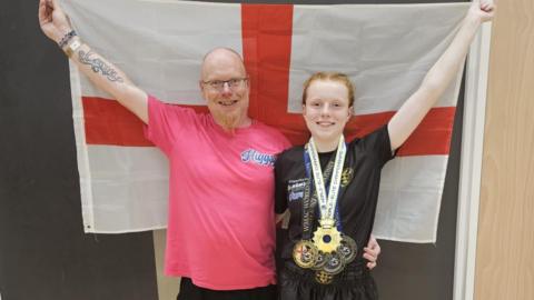 Alan wearing a pink T-shirt standing next to Lana wearing a black T-shirt and medals around her neck - with both of them holding a St George's flag behind them.
