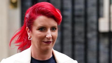 Louise Haigh appearing outside the front of Downing Street. She has distinctive dyed red hair, and is wearing a cream blazer and blue top. 