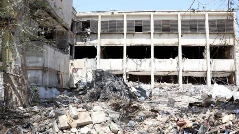 A building at Evin prison is shown damaged. A three-storey white building is seen standing, its windows blown out by an explosion. Extensive rubble is seen in fron tof the building, with piles of concrete and metal shown. A tree branch can be seen at the edge of the photograph.