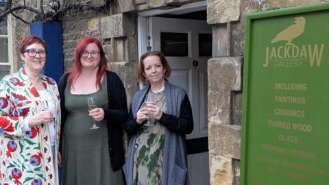 Three women standing outside a stone doorway beneath a green ‘Jackdaw Gallery’ sign, each holding a drink; the gallery entrance and signage are visible behind them.