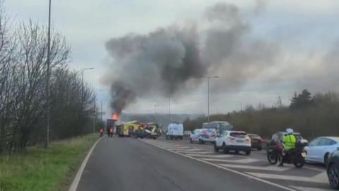 A fire crew uses a high-powered hose to dampen down a smoking lorry, which was on fire. There is a fire engine in the foreground.