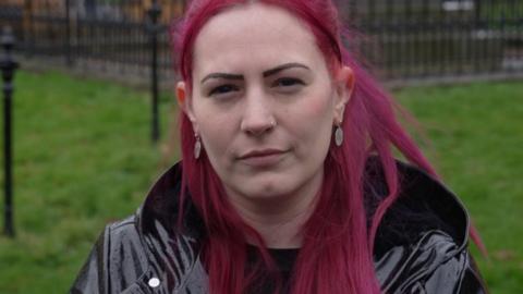 A woman with red hair, staring at the camera, wearing a dark shiny coat