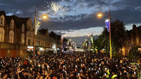 Crowds of people gather in the road as fireworks light up the sky