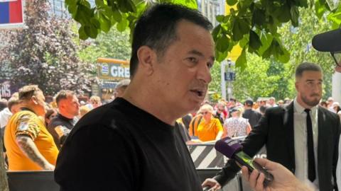 Hull City owner Acun Ilicali being interviewed outside Wembley Stadium, wearing a black t-shirt. Crowds on Olympic Way can be seen behind him.