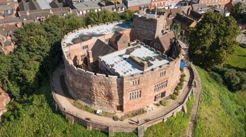 An aerial view of Tamworth Castle on a sunny day, where its grassed mount has residential and industrial buildings behind it.