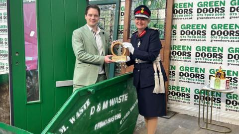 Joseph Holman is standing in a green skip with a green door placed behind him, also in the skip. He is wearing a mint-coloured suit as he receives the King's Award for Enterprise: Sustainable Development from Susan Lousada, the Lord-Lieutenant of Bedfordshire. She is showing off her distinctive regalia. 