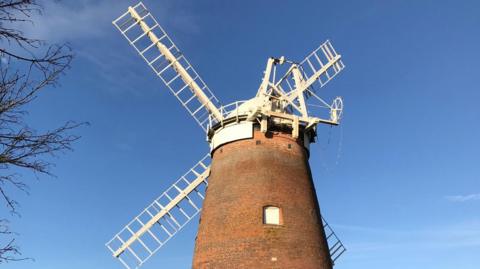 A brick windmill with four white sails pictured on a sunny day