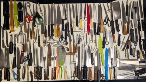 A large assortment of kitchen knives and utensils is neatly arranged on a white surface. The collection includes chef’s knives, cleavers, serrated knives, paring knives, utility knives, and multiple pairs of scissors. Also visible are sharpening steels, peelers, can openers, ice cream scoops and spoons.