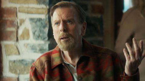 Timothy Spall as John Chapel in Death Valley. He is wearing a colourful checked shirt and looking chastened. Exposed brickwork is visible behind.