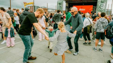 A boy and girl dancing with two men. The boy has his face painted. A large crowd and a stage are visible in the background. One of Harland & Wolff's yellow cranes is to the side of the grounds. 