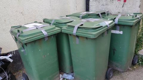 Five green bins in an alleyway, taped up with grey tape