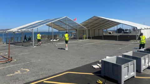 The car park space has two large marquee structures being built, one has a white covering over its roof and two sides. Three people in hi-vis jackets work on the site. You can see the sea in the distance.