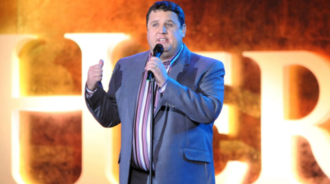 Peter Kay performing on stage in a blue suit and stripy shirt