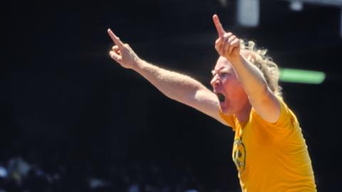 An old man in a yellow shirt and denim shorts raises his index fingers in the air at a sporting event.