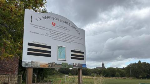 A sign which reads T.V. Harrison Sports Ground with a football field in the background.