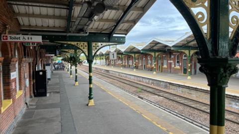 Two platforms on a Victorian railway station have ornate yellow and green pillars and arches made of iron. There are modern-day signs for passengers and train tracks between the two platforms