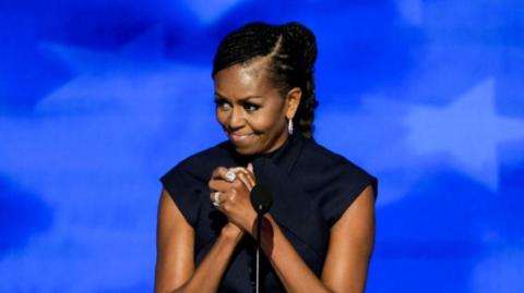 Michelle Obama stands on stage with her hands clasped together and a smile on her face at the Democratic National Convention in Chicago