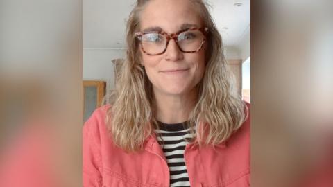 A woman, Helen Upshall, wears a striped tshirt with orange shirt over the top and glasses. She has long blonde hair.