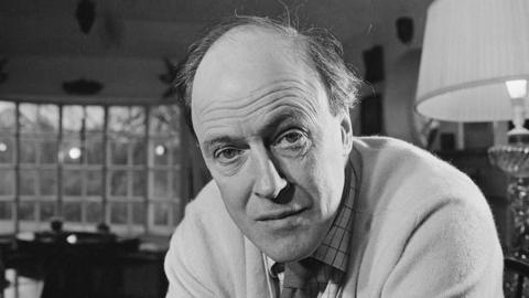 Welsh writer Roald Dahl looks at the camera. The photo is in black and white. He has dark brown hair and is balding on the top of his head. Dahl is wearing a light-coloured knit, white check shirt and dark tie. 